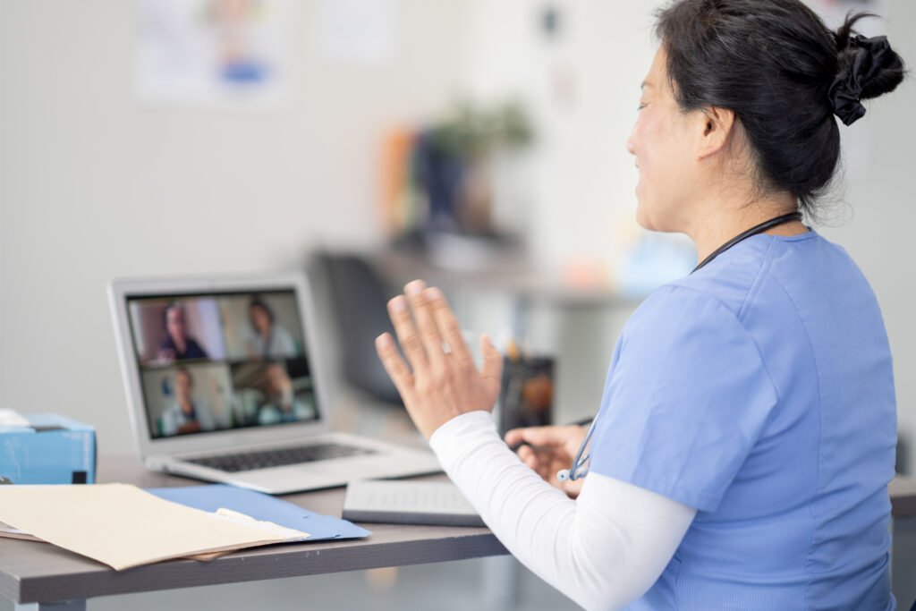 A female doctor of Asian decent, sits in front of her computer as she participates in a virtual meeting with four other medical professionals.  She is wearing blue scrubs and has her back towards the camera as she talks with her colleagues on the screen.