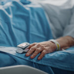 Hospital Ward: Senior Woman Resting in a bed with Finger Heart Rate Monitor / Pulse oximeter showing Pulse. Her Fragile Hands Resting on a Blanket. Focus on the Hand.