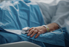 Hospital Ward: Senior Woman Resting in a bed with Finger Heart Rate Monitor / Pulse oximeter showing Pulse. Her Fragile Hands Resting on a Blanket. Focus on the Hand.