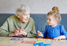 Beautiful toddler girl and grand grandmother playing together pictures lotto table cards game at home. Cute child and senior woman having fun together. Happy family indoors.