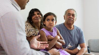 Mature Aboriginal couple sit with their cute granddaughter on their lap while a multiracial healthcare professional talks to them in a waiting room