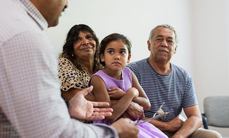 Mature Aboriginal couple sit with their cute granddaughter on their lap while a multiracial healthcare professional talks to them in a waiting room