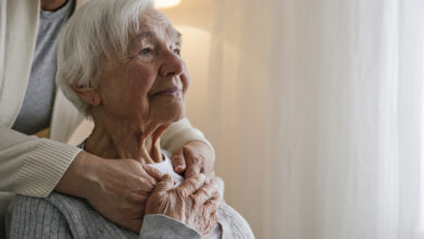 Unrecognizable female expressing care towards an elderly lady, hugging her from behind holding hands. Two adult women of different age. Family values concept. lose up, copy space, background.
