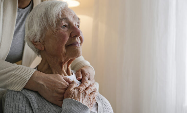 Unrecognizable female expressing care towards an elderly lady, hugging her from behind holding hands. Two adult women of different age. Family values concept. lose up, copy space, background.