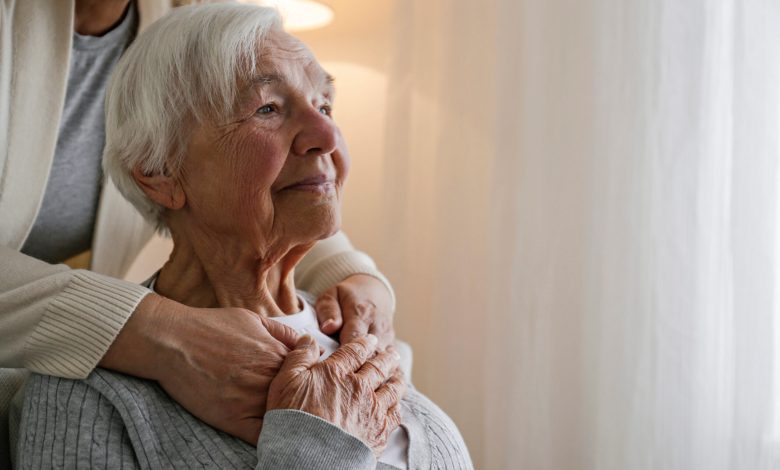Unrecognizable female expressing care towards an elderly lady, hugging her from behind holding hands. Two adult women of different age. Family values concept. lose up, copy space, background.