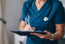 A close-up of a healthcare professional in blue scrubs writing notes on a clipboard, symbolizing care and medical professionalism in a modern medical setting.