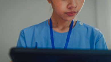 Closeup of young Asian female nurse in blue scrubs sitting on bed while using a digital tablet. Nurse lifestyle concept.