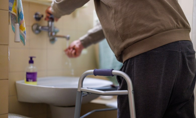 An older individual uses a sink and soap to wash their hands, with a walker positioned close for stability and assistance