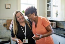 Waist-up view of Black client and healthcare worker standing with arms around each other and laughing during routine house call.