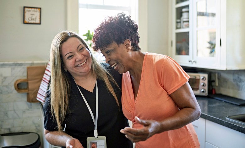 Waist-up view of Black client and healthcare worker standing with arms around each other and laughing during routine house call.
