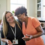 Waist-up view of Black client and healthcare worker standing with arms around each other and laughing during routine house call.