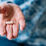 This close-up photograph features a 101 year old woman holding prescription medication pills and Vitamin Supplements in her hand. She has a bandage on her thumb. The senior adult is seated in the comfort of her domestic living room. Handling and taking pills or capsules can be difficult for individuals suffering from arthritis. The photograph captures the real-life choices and healthcare practices of this one woman as she has crossed the milestone of living beyond 100 years. The image shows the importance of medicine and healthcare in maintaining a long and healthy life.