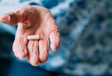 This close-up photograph features a 101 year old woman holding prescription medication pills and Vitamin Supplements in her hand. She has a bandage on her thumb. The senior adult is seated in the comfort of her domestic living room. Handling and taking pills or capsules can be difficult for individuals suffering from arthritis. The photograph captures the real-life choices and healthcare practices of this one woman as she has crossed the milestone of living beyond 100 years. The image shows the importance of medicine and healthcare in maintaining a long and healthy life.