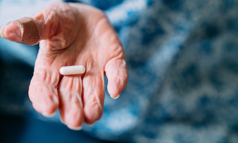 This close-up photograph features a 101 year old woman holding prescription medication pills and Vitamin Supplements in her hand. She has a bandage on her thumb. The senior adult is seated in the comfort of her domestic living room. Handling and taking pills or capsules can be difficult for individuals suffering from arthritis. The photograph captures the real-life choices and healthcare practices of this one woman as she has crossed the milestone of living beyond 100 years. The image shows the importance of medicine and healthcare in maintaining a long and healthy life.