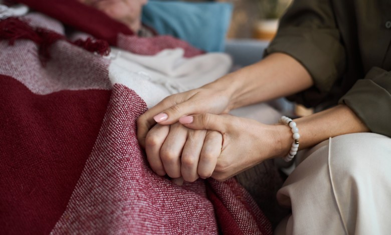 Middle aged Caucasian woman holding hand of senior Caucasian man lying under blanket, showing support and comfort, close up on hands emphasizing caregiving and compassion