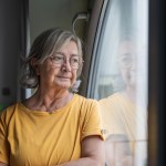 Senior woman stands by a window, gazing outward in quiet reflection; contemplative, solitary moment capturing aging, memory, hope and the calm dignity of later life at home
