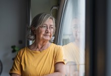 Senior woman stands by a window, gazing outward in quiet reflection; contemplative, solitary moment capturing aging, memory, hope and the calm dignity of later life at home