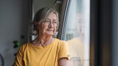 Senior woman stands by a window, gazing outward in quiet reflection; contemplative, solitary moment capturing aging, memory, hope and the calm dignity of later life at home