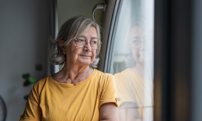 Senior woman stands by a window, gazing outward in quiet reflection; contemplative, solitary moment capturing aging, memory, hope and the calm dignity of later life at home