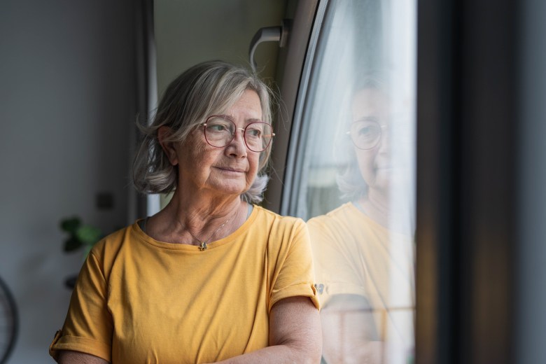 Senior woman stands by a window, gazing outward in quiet reflection; contemplative, solitary moment capturing aging, memory, hope and the calm dignity of later life at home
