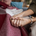 Middle aged Caucasian woman holding hand of senior Caucasian man lying under blanket, showing support and comfort, close up on hands emphasizing caregiving and compassion