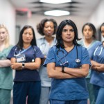 Portrait of a team of female nurses and doctors. The multi-ethnic medical professionals are standing in a corridor of a hospital. The confident group is looking directly at the camera. They have serious expressions on their faces and their arms are crossed. A doctor of Indian descent is standing at the front of the group.