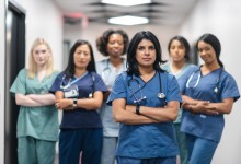 Portrait of a team of female nurses and doctors. The multi-ethnic medical professionals are standing in a corridor of a hospital. The confident group is looking directly at the camera. They have serious expressions on their faces and their arms are crossed. A doctor of Indian descent is standing at the front of the group.