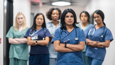 Portrait of a team of female nurses and doctors. The multi-ethnic medical professionals are standing in a corridor of a hospital. The confident group is looking directly at the camera. They have serious expressions on their faces and their arms are crossed. A doctor of Indian descent is standing at the front of the group.