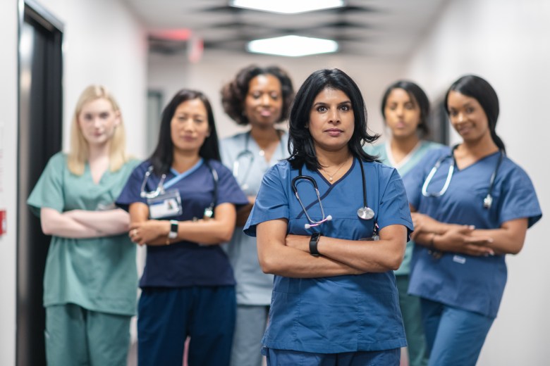 Portrait of a team of female nurses and doctors. The multi-ethnic medical professionals are standing in a corridor of a hospital. The confident group is looking directly at the camera. They have serious expressions on their faces and their arms are crossed. A doctor of Indian descent is standing at the front of the group.