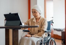 A senior Asian female cancer patient is sitting in a wheelchair and having a video call with her doctor using a laptop in the living room.