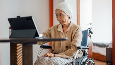 A senior Asian female cancer patient is sitting in a wheelchair and having a video call with her doctor using a laptop in the living room.