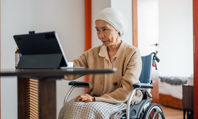 A senior Asian female cancer patient is sitting in a wheelchair and having a video call with her doctor using a laptop in the living room.