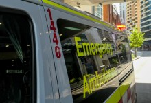 An ambulance of the New South Wales Ambulance Service parked on Oxford Street, Bondi Junction, Sydney. The emergency red light is flashing on the top of the rear door. In the background are commercial and residential buildings. This image was taken on a sunny morning on 11 December 2023.