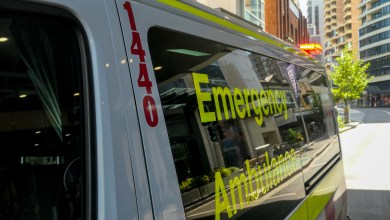 An ambulance of the New South Wales Ambulance Service parked on Oxford Street, Bondi Junction, Sydney. The emergency red light is flashing on the top of the rear door. In the background are commercial and residential buildings. This image was taken on a sunny morning on 11 December 2023.